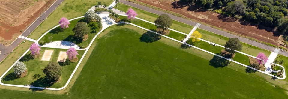 Vista aérea da praça: gramado amplo, caminhos claros em curva, duas quadras de areia, ipês floridos e a mata ao fundo